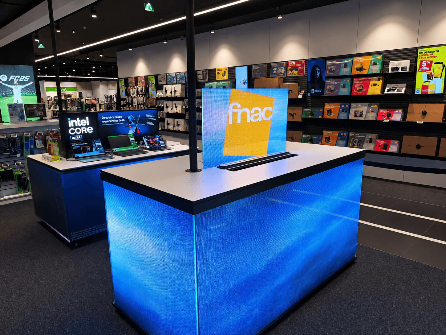 Balcão azul iluminado com logótipo Fnac em loja de eletrónica com computadores portáteis em exposição.