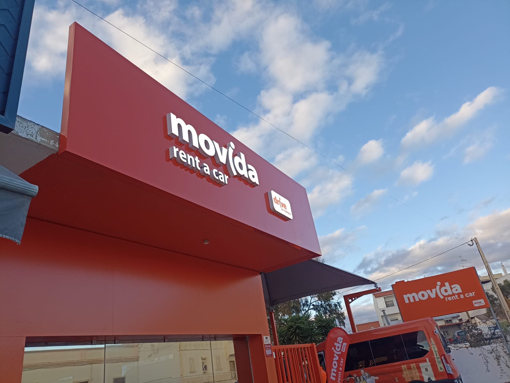 Fachada vermelha da Movida Rent a Car sob céu azul com nuvens esparsas.